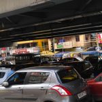 A congested street scene underneath an overpass, filled with various cars. Behind the vehicles, there are several shops with colorful signs and billboards, including one for "Kalyan Jewellers" and another for "SKSE Jewellers." People are walking on the sidewalks.