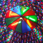 A vibrant, colorful dome covered in a mosaic of red, blue, green, and purple triangular patterns with varied lighting effects. The roof features hanging light fixtures that resemble bottles, adding to the kaleidoscopic appearance.