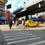 A busy urban street scene shows pedestrians crossing at a crosswalk, with a yellow taxi driving through the intersection. The background features various shops, advertisements, and an overhead bridge.