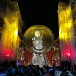 A large crowd gathers at night to view a towering elaborate statue adorned with lights and decorations during a festival. The statue is flanked by illuminated structures with yellow and red hues, creating a vibrant and festive atmosphere.