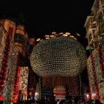 A large circular lantern decorated with numerous small lights is suspended in the air amidst a vibrant festival. The area is adorned with intricate patterns and bright colors. Crowds of people gather below, and buildings are visible in the background under the night sky.