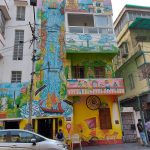 A vibrant building façade with intricate, colorful murals depicting various scenes, including a flowing river, sailing ships, and landscapes. The artwork extends across multiple stories. A car is parked in front of the building, and some people are nearby.