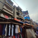 A street market selling clothes is set up in front of a series of multi-story buildings. The buildings have balconies with railings, some with laundry hanging. Overhead wires run across the scene, and a cloudy sky is in the background.