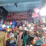 A busy market stall with a sign reading "M.S. Chowdhury Jewellers." The stall is adorned with various bags, backpacks, and merchandise. People are browsing and inspecting items. The atmosphere appears lively with bright lighting and a crowded space.