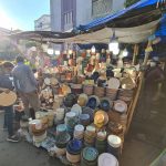 A street market stall displays a wide variety of ceramic dishes, including plates, bowls, and cups in different colors and styles. Shoppers browse the selection under a canopy of bright lights, with buildings visible in the background.