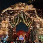 People gather at night to admire a large, intricately decorated festival archway. The arch is adorned with colorful lights and various materials, and a vibrant, illuminated design is visible through the arch. Tree branches frame the structure from above.