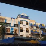 The image shows a large, multi-story building with white and blue paint. The structure displays multiple signs with "LIC" in bold letters. Several small shops with blue tarpaulin-covered roofs are in front of the building. Palm trees are seen around the area.