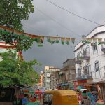 A street scene under a cloudy sky with buildings on both sides. There are colorful flags strung overhead, a yellow auto-rickshaw in the foreground, and various market stalls and people walking around. Brightly painted buildings contrast against the dark clouds.