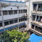 A view of an old, multi-story building with worn, white paint and numerous windows, partly covered by rusted grilles. In the foreground, there's a small courtyard with a green leafy tree and a large blue tarpaulin. The sky above is clear and sunny.