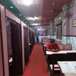 A narrow, dimly lit restaurant with red and white ceiling decor. Booths with curtains line one side, white and pink walls the other. A man in a red shirt sits at a table on the right, looking at his phone. Another person stands in the background near the entrance.