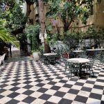 A quaint outdoor café features a black and white checkered floor, several round tables with wrought iron chairs, and lush greenery surrounding the seating area. A pathway lined with plants leads into the distance, creating a serene and inviting atmosphere.