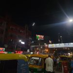 Nighttime street scene bustling with people, auto rickshaws, and colorful neon signs. A flyover bridge and billboard can be seen in the background with a mix of commercial buildings and street vendors completing the lively urban setting.