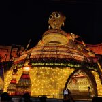 A nighttime scene of a large, brightly illuminated decorative structure resembling an intricate art installation. The structure features a dome and a large humanoid figure on top, draped in colorful lights. Silhouetted people and someone taking a photo are visible in the foreground.