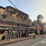 A street scene in an urban area with weathered buildings and various shops. People walk along the sidewalk, with signs in both English and Bengali above the storefronts. Power lines crisscross the sky, and a "STOP" marking is visible on the road.