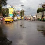 A busy urban street scene after rainfall, featuring yellow taxis, green auto-rickshaws, and pedestrians on a wet road. Buildings with billboards and trees line the sides of the street. The sky is overcast with gray clouds.