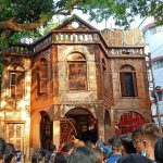 A weathered, two-story building with arched windows and wooden shutters stands under the shade of a large tree. A crowd of people, many holding up their phones, gathers in front. The scene is set in an urban area with modern buildings in the background.