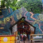 A bustling crowd walks toward a large, intricately decorated structure with futuristic circuit board patterns at the entrance. The colorful design, with abstract shapes and lines, contrasts against the greenery and buildings surrounding it. A "Kolkata Traffic Police" barricade is visible.