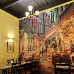 A cozy restaurant interior with yellow walls and a mural depicting a lively Parisian street scene with outdoor cafes and colorful signs. Wood tables and chairs are set for diners, and a chandelier hangs from the ceiling. A framed picture adorns one wall.