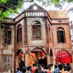 People gather in front of a large, rustic, and intricately designed wooden structure with multiple windows and arches. Inside, a brightly colored idol of a deity is displayed under a canopy of vibrant red and orange drapery. Trees and surrounding buildings are visible.