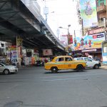 A bustling city intersection with various cars including a yellow taxi and a blue bus under a flyover. The streets are lined with buildings adorned with numerous billboards and advertisements. Pedestrians are visible walking along the sidewalks.