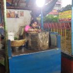 A woman is cooking at a blue street food stall under a canopy. She is preparing food in a large metal pot, surrounded by various cookware. In the background are colorful posters, a yellow sign, and a busy street with a fence. The scene is evening or night.