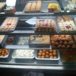 A display case in a bakery showing a variety of sweets and desserts, including round pastries, square pieces, and rolled items, arranged on trays and plates. Each shelf is filled with an assortment of colorful and differently shaped treats.