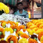 A flower vendor in a market sits behind an array of colorful flowers, including yellow and orange marigolds, white jasmine, and other blossoms. He is wearing a striped shirt, and the background features hanging items and dim lighting.