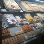 A display case filled with a variety of Indian sweets, including stacked trays of different types of burfi, peda, and other desserts. Each tray has a red card with a price tag. The case is well-lit, and all sweets are neatly arranged.