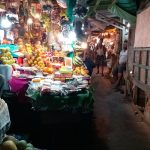 A dimly lit market aisle with various stalls displaying fruits, vegetables, and other produce. The stall in the foreground has bright lights illuminating the colorful fruits. People are seen walking and standing further down the aisle.