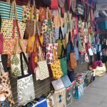 A sidewalk shop displays a colorful variety of tote bags hung in rows on a wall. The bags feature different patterns such as floral, striped, and geometric designs. The ground also has additional bags, and the shop's metal shutter is visible behind the display.