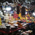 A bustling market stall displays a colorful assortment of fruits, vegetables, dried goods, and packaged food products. The well-lit stall features various items such as oranges, apples, pumpkins, spices, and jars of preserves. Shoppers are visible in the background.