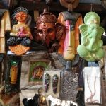 A vibrant market stall displays an array of colorful, handcrafted masks and statues. Among them are masks of deities, including Ganesha and Buddha. Beaded necklaces and other decorative items hang overhead, adding to the rich, artistic ambiance.