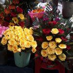 A vibrant flower shop display featuring assorted bouquets. Yellow roses dominate the center in a bucket, surrounded by roses of pink and red hues. White flowers and additional floral arrangements are visible, with plastic stools and crates holding the display.