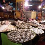 A bustling seafood market stall displaying a variety of fresh seafood, including fish, shrimp, and squid, arranged on trays. Two vendors are visible in the background, and the stall is illuminated by hanging light bulbs, creating a lively and busy atmosphere.