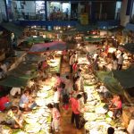 A bustling indoor fish market with numerous vendors displaying a variety of seafood on large trays. Shoppers move through the narrow aisles, examining the fresh catch under bright lights. Green tarps hang overhead, and multi-level shops are visible in the background.