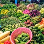 A vibrant display of various fresh vegetables, including carrots, cucumbers, beans, bell peppers, eggplants, pumpkins, green peas, and cluster beans, arranged in piles at a market. A pink basket filled with cucumbers sits in the foreground.