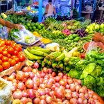 A vibrant market stall filled with a variety of fresh vegetables and fruits including tomatoes, onions, bananas, cucumbers, and eggplants. Two vendors in white tank tops are busy sorting and bagging produce for customers under bright lighting.