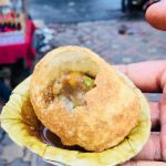 Close-up of a hand holding a single serving of a filled pani puri on a leaf plate. The pani puri has a visible bite taken out, revealing its potato and chickpea filling. The background shows a slightly blurred street scene with a few people and vendors.