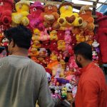 Two men stand in front of a street vendor's stall filled with colorful stuffed animals, including teddy bears and other plush toys, in various sizes and colors. The men appear to be browsing the toys. The background shows part of the outdoor market setup.