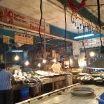 A fish market with vendors and various types of fish displayed on tables under bright hanging lights. One vendor is wearing a yellow shirt and is attending to the fish, while another person in a checkered shirt stands by. The area is cluttered with posters and signs.