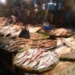 A bustling seafood market stall displays a variety of fresh fish, prawns, and other seafood on ice and banana leaves under bright lights. Two vendors are seated in the background, one inspecting a fish, with the market appearing busy and vibrant.