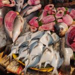 A market stall displays various types of fresh fish, including whole fish and fish cut into pieces. Some fish are arranged on ice, while others rest on green leaves. The display includes both shiny silver fish and fish with darker scales and pink flesh.