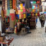 A bustling street market with vibrant, patterned fabrics hanging on display. Several people sit and stand near the shop, which has colorful lights and a cloth canopy. The ground is paved with interlocking stones, and other market stalls are visible in the background.