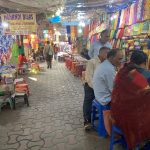 A bustling market scene with colorful fabric stalls on both sides. People are seated on small stools near the stalls, engaged in conversation or waiting. The ceiling is covered with plastic sheets, and a sign reading "MEHANDI BILAS" is visible in the background.