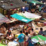 An outdoor market scene with various stalls covered by tarps. Several vendors and shoppers are interacting amidst the vibrant and busy environment. Fresh produce and goods are displayed on tables. The area is bustling with activity and illuminated by hanging lights.