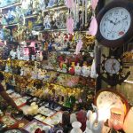 A crowded shop display filled with a variety of decorative items including clocks, porcelain figurines, vases, candles, and other trinkets. Shelves are packed with colorful and intricate ornaments, creating a visually rich and eclectic scene.