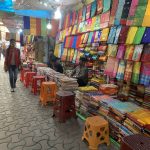 A colorful fabric shop with vibrant cloths displayed on the walls and stacks of folded fabrics on tables. Three sellers are seated on stools, attending to customers. The pavement is lined with stools, and a few people walk by. The shop is covered with a transparent canopy.