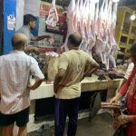 Three men are standing in front of a butcher's stall in a market, observing the meat. The butcher is behind the counter, cutting pieces of meat, with several large hanging cuts of meat in the background. The surroundings look busy with hanging lights above the stall.