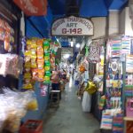 A narrow, busy market alley lined with colorful snack packets, household items, and various goods hanging on the walls. People are seen shopping and walking down the aisle. A sign above reads "Robin Thermocol Art G-142." The scene is vibrant and bustling.