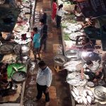 Bird's-eye view of a bustling fish market at night. Several vendors display fresh fish on tables illuminated by bright lamps. Corridors are wet and crowded with people buying and selling seafood. Various fish are arranged on beds of ice and in metal trays.